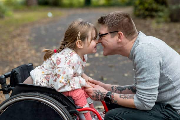 A close up side view of a father and his young daughter who is a wheelchair user having a cute affectionate moment with each other while out in a beautiful park